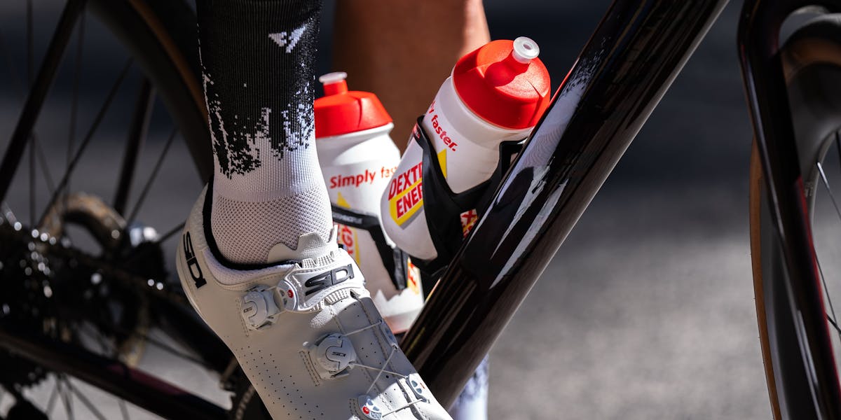 Cyclist's shoe on pedal with energy drink bottles attached to bike frame.