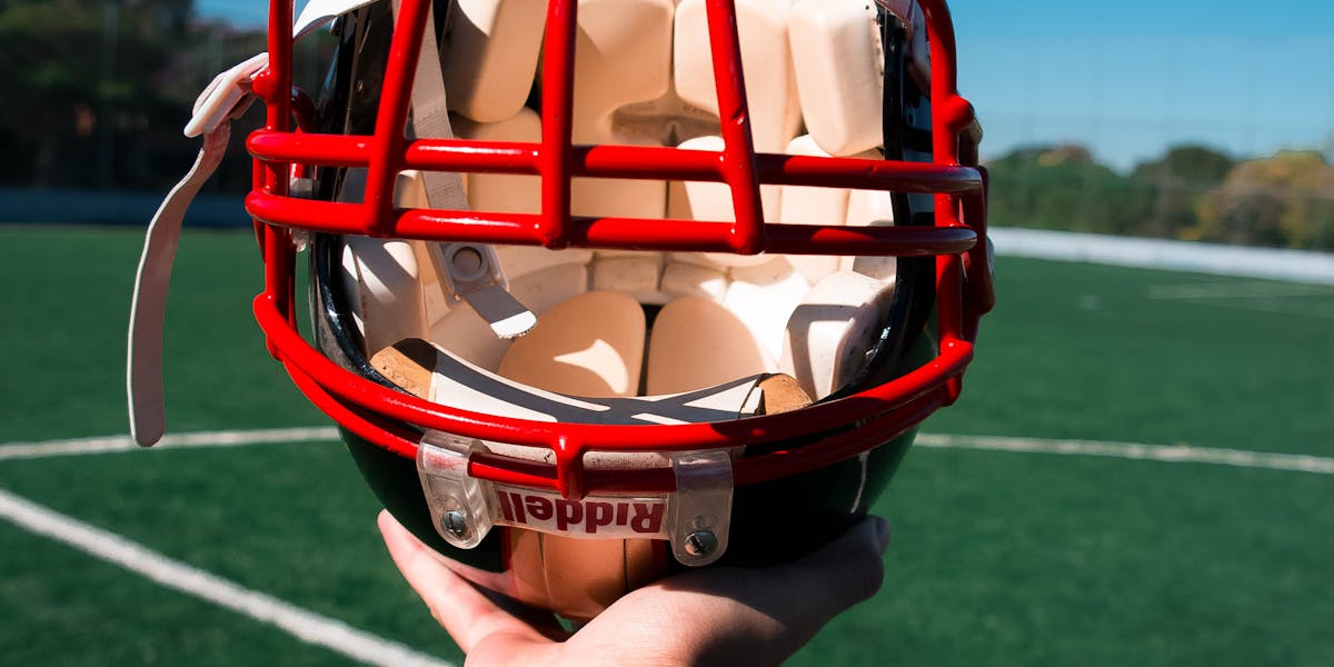 Close-up of a football helmet held by tattooed arm on a sunny sports field.