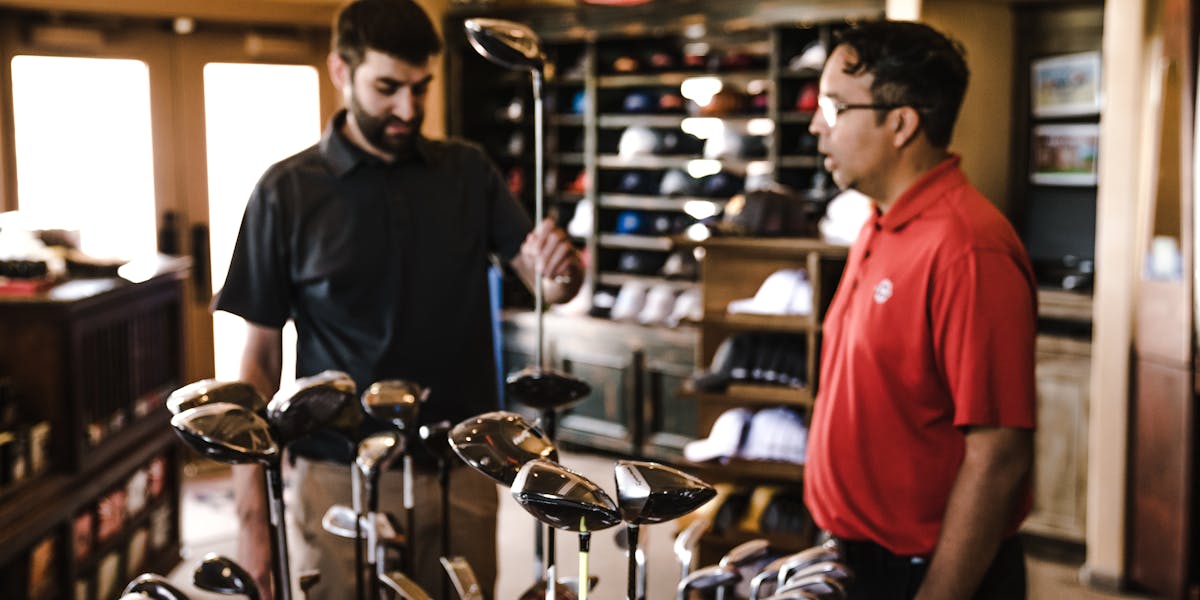 Two men browsing golf clubs in a sports equipment store, discussing options.