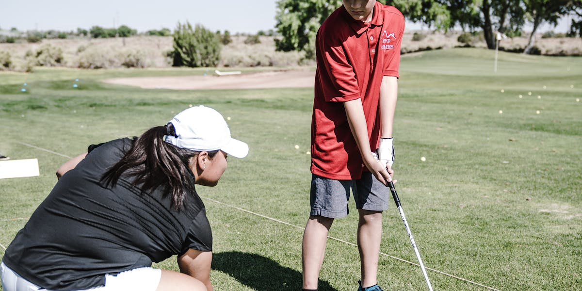 A golf instructor guides a young golfer on the green during a sunny practice session.