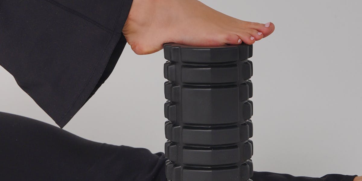 Person using a black foam roller for leg exercise on a gray background.