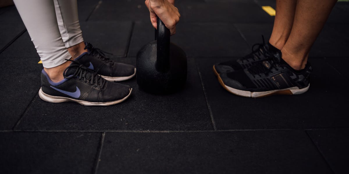Close-up of two people lifting a kettlebell in a gym setting, emphasizing footwear...