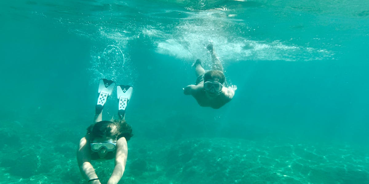 Two adults snorkeling underwater in a clear blue sea with fins and masks.