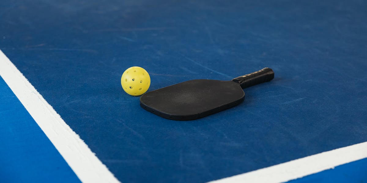 A pickleball paddle and yellow ball on a blue court corner, symbolizing sport an