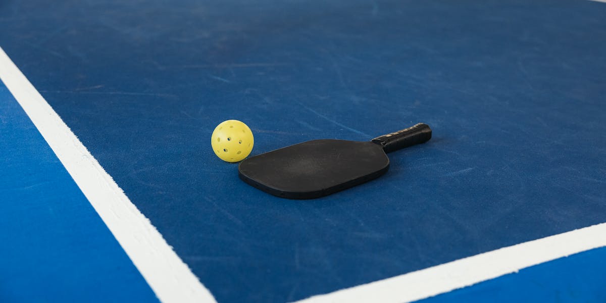 A pickleball paddle and yellow ball resting on a blue pickleball court with white ...