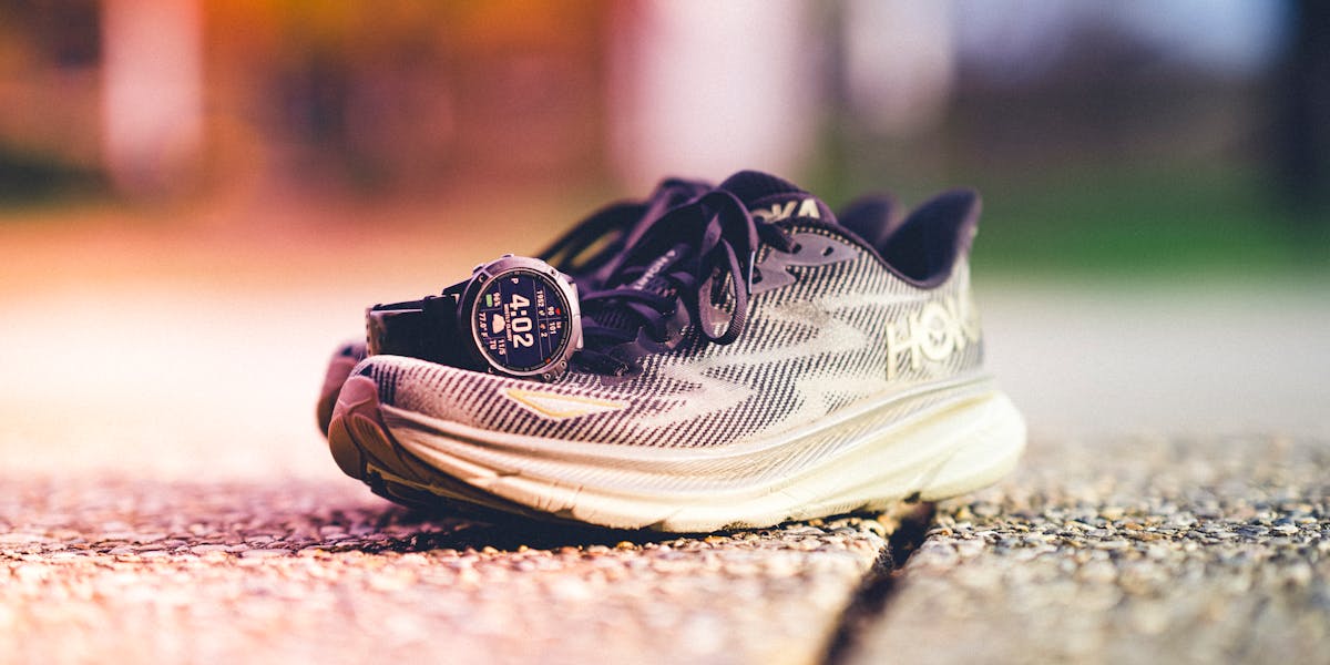 A pair of Hoka running shoes with a Garmin watch beside them on a sunny outdoor tr...