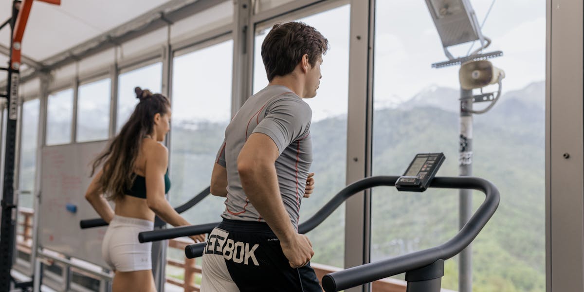 Two fitness enthusiasts running on treadmills in a gym with outdoor view.