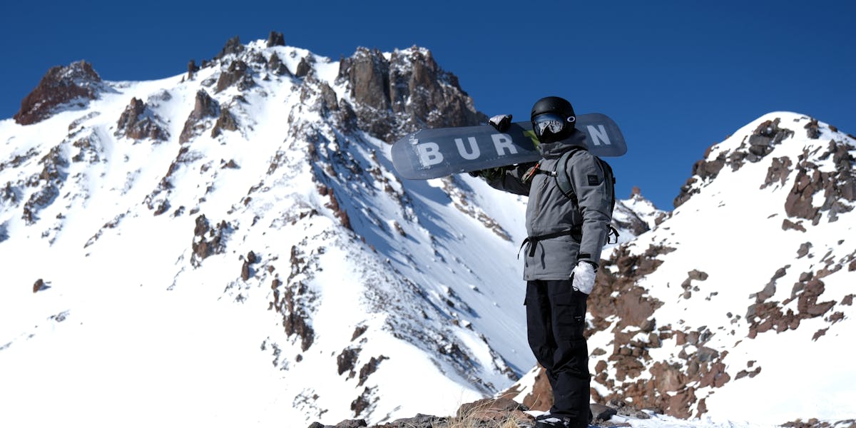Snowboarder standing on snowy mountain peak holding Burton board under clear blue ...