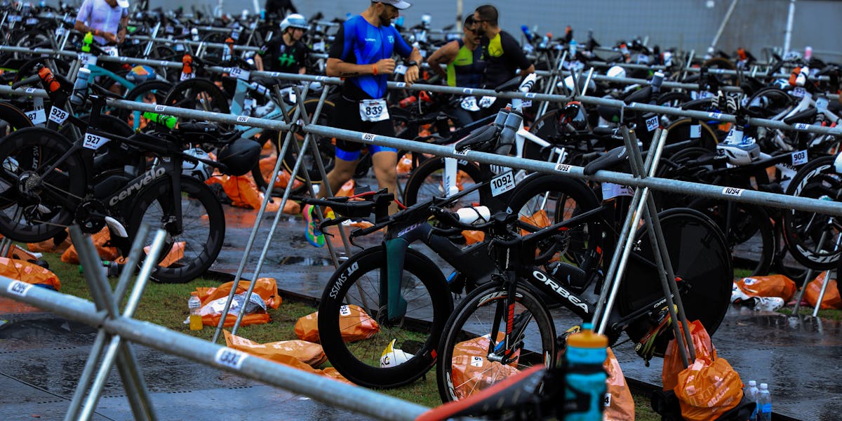 Athletes in a triathlon transition zone surrounded by multiple bicycles and gear