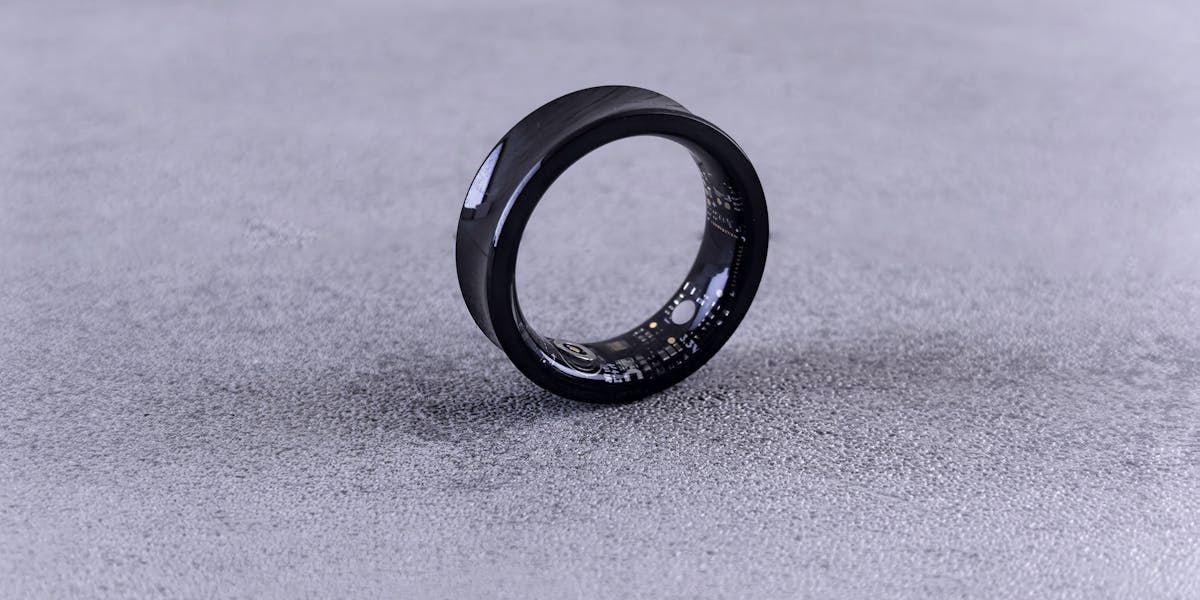 A modern black smart ring with visible circuitry resting on a textured concrete su...
