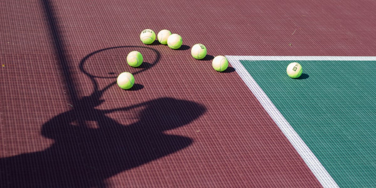 A tennis racket shadow and scattered balls on a colorful court under sunlight.