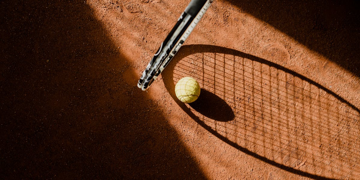 Tennis ball with racket shadow on a sunlit clay court.